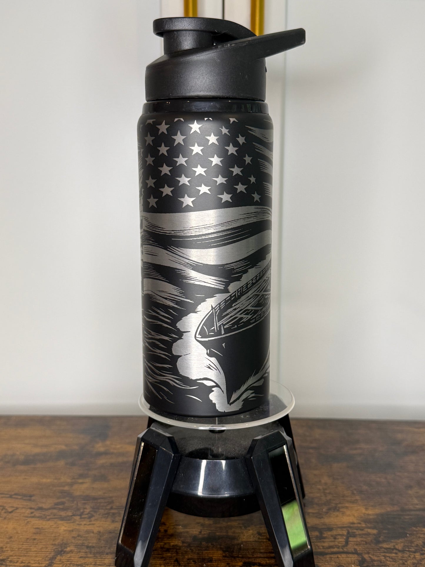A black insulated water bottle with a flip-top lid, featuring a silver and black American flag design with a running dog graphic, displayed on a clear stand on a wooden surface.