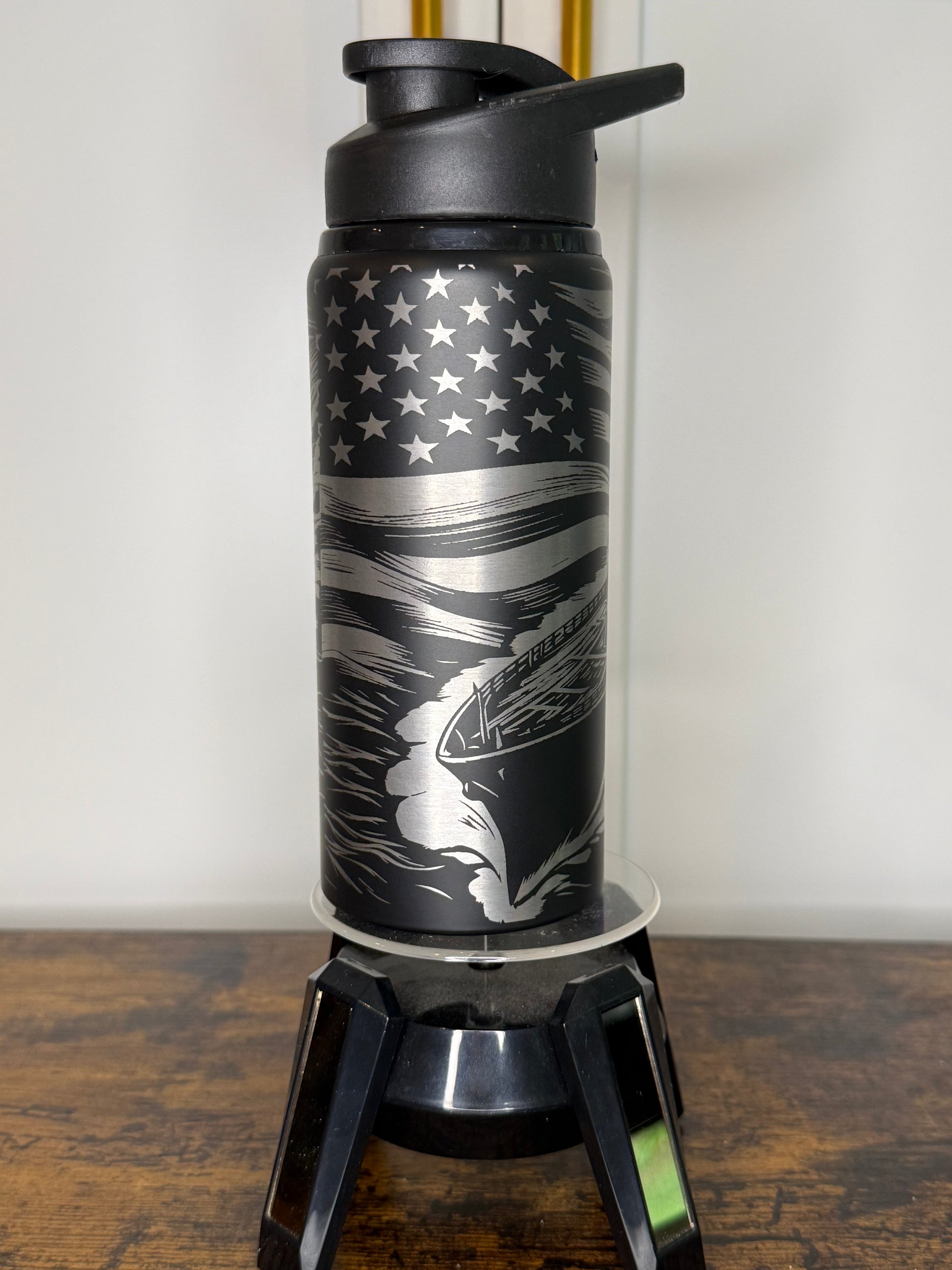 A black insulated water bottle with a flip-top lid, featuring a silver and black American flag design with a running dog graphic, displayed on a clear stand on a wooden surface.