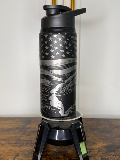 A black insulated water bottle with a flip-top lid, featuring a silver and black American flag design with a running dog graphic, displayed on a clear stand on a wooden surface.