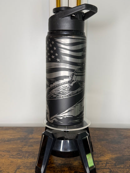 A black insulated travel tumbler with a matching black lid and handle, featuring a silver etched American flag and ship design, displayed on a clear stand on a wooden surface.